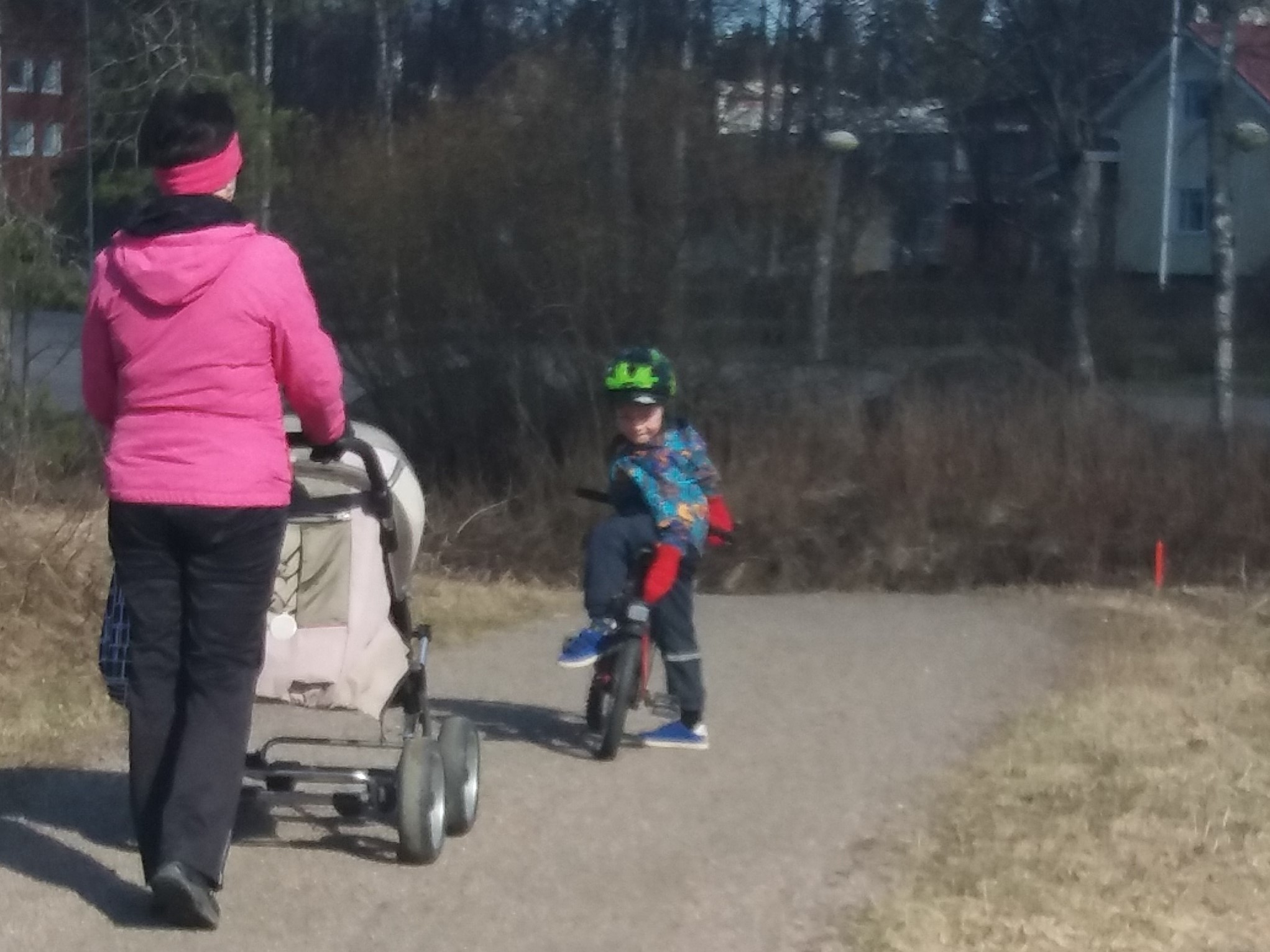 "Nainen työntää lastenvaunuja. Leikki-ikäinen lapsi odottaa pyörän kanssa edellä."